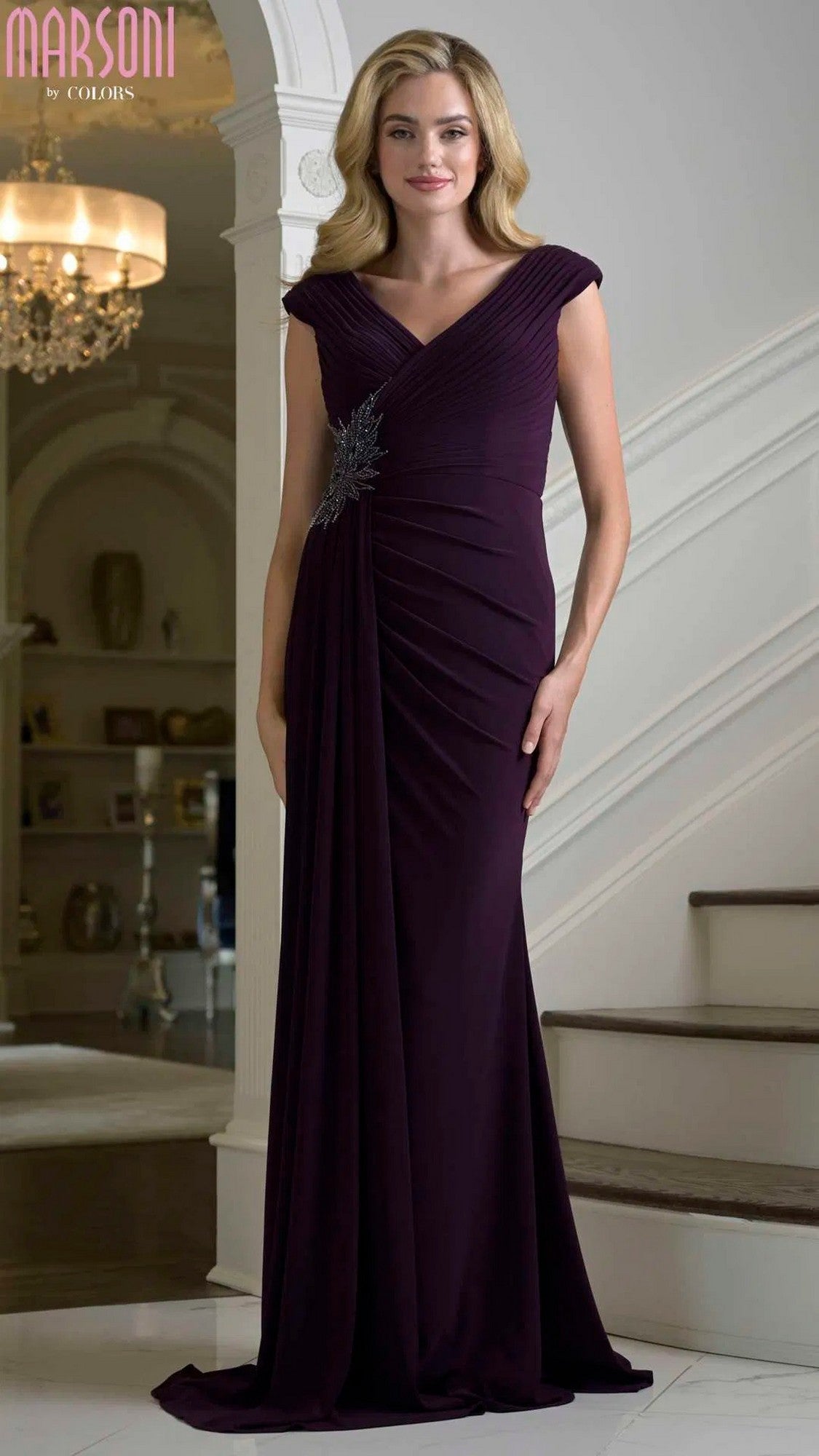 A woman stands indoors wearing the Marsoni M343 long formal dress in eggplant, featuring a ruched fitted bodice, cap sleeves, and a silver embellishment at the waist.